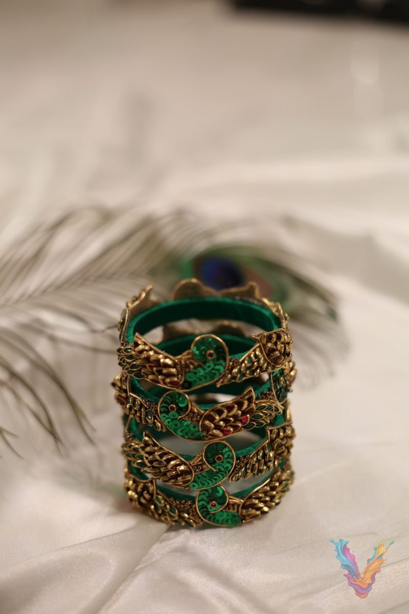 A stack of green silk thread bangles with gold peacock embellishments on white satin